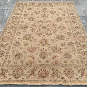 Könnte beinhalten: Beiger rechteckiger Teppich mit einem aufwendigen Blumenmuster. Der Teppich hat ein zentrales Feld mit sich wiederholenden floralen Motiven und einen detaillierten Rand. Die Farbpalette umfasst Beige, Braun und rote Akzente, was eine warme Ästhetik schafft.