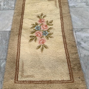 May include: A rectangular rug with a textured, beige pile surface featuring a central floral motif of pink roses and blue flowers with green leaves. The rug is framed by a thin, dark brown rectangular border and a wider, textured beige border.