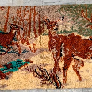 May include: A rectangular rug featuring a forest scene with deer. The rug has a beige background with brown, green, and teal accents. Two deer are in the foreground, with a group of deer in the background. The rug is made of a textured material.