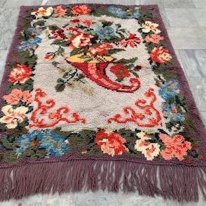 May include: Rectangular rug with a floral design. The rug features a central cornucopia filled with flowers in shades of red, orange, and yellow. The border is a deep purple with fringe.