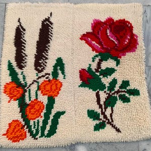 May include: A cream-colored, textured rug with embroidered floral designs. One side features orange and brown stylized flowers and foliage. The other side displays a large red rose with green leaves and stems. The rug has a soft, plush appearance.
