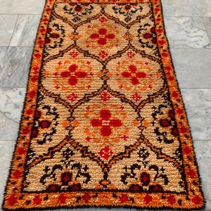 May include: Rectangular rug with a repeating floral pattern in shades of beige, orange, red, and black. The rug has a textured, woven appearance and a fringed border. The design features stylized floral motifs.