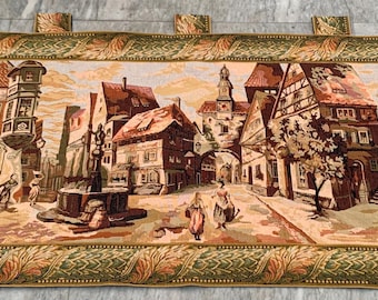 Könnte beinhalten: Ein Wandteppich, der eine europäische Stadtszene mit Gebäuden, Menschen und einem Brunnen darstellt. Der Wandteppich hat einen detaillierten Rand mit einem goldenen und grünen Design. Die Farbpalette umfasst Braun-, Beige- und Goldtöne.