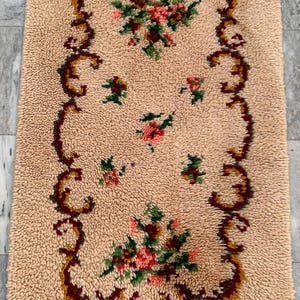 May include: A rectangular beige shag rug featuring a central floral motif with pink and red flowers and green leaves. The design is enclosed by a dark red and brown scrolled border, creating a decorative frame on the textured pile surface.