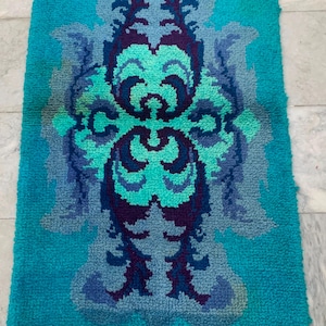 May include: A rectangular, turquoise rug with a central, ornate design in shades of blue, teal, and purple. The rug has a textured, looped pile and a symmetrical, floral-like pattern. The rug is approximately 36 inches long and 24 inches wide.