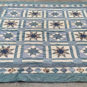 Op de afbeelding: Een grote, vierkante quilt met een blauw en wit sterrenpatroon. De quilt heeft een centraal raster van sterrenpatronen in blauw- en crèmetinten, omlijst door een patroonrand. De buitenrand is effen blauwgroen.