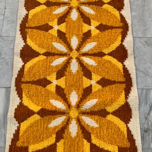 May include: A rectangular rug with a repeating floral pattern in shades of yellow, brown, and white. The rug has a cream-colored background and a textured, woven appearance. The floral design is symmetrical and stylized.
