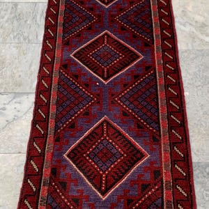 May include: A rectangular, hand-woven rug with a geometric pattern in shades of red, burgundy, and purple. The rug features diamond and triangular shapes, with a fringed edge. The rug is placed on a marble surface.