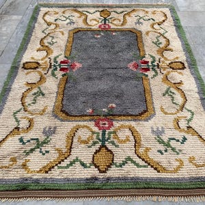 May include: Rectangular rug with a central gray field framed by a cream-colored border. The border features floral and scroll designs in gold, green, and red. The rug has a green border and a fringed edge.