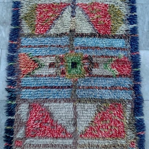 May include: Rectangular, hand-woven rug with a geometric pattern. The rug features a mix of colors, including red, blue, green, and white. The design includes triangles and horizontal stripes. The rug has a dark blue fringe around the edges.