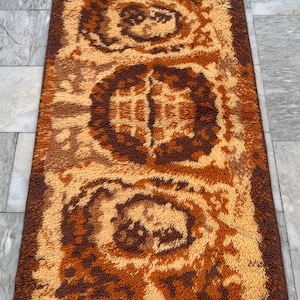 May include: Rectangular shag rug with a repeating pattern in shades of brown and tan. The rug features a dark brown border and a central design of concentric circles and geometric shapes. The texture appears soft and plush.