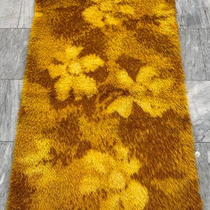 May include: A rectangular, shaggy rug in shades of gold and brown. The rug features a floral pattern in a lighter gold hue against a darker brown background. The rug's texture appears soft and plush, suitable for a living room or bedroom.