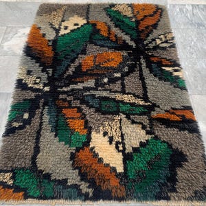 May include: A rectangular rug with a shaggy texture. The rug features a leaf-like pattern in shades of brown, green, gray, and cream against a dark background. The rug's design is abstract and textured, suitable for home decor.