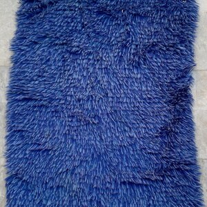May include: A rectangular, shaggy rug in a vibrant shade of blue. The rug has a thick, textured pile, giving it a plush appearance. The edges are finished with a cream-colored trim. This rug would be a cozy addition to any room.