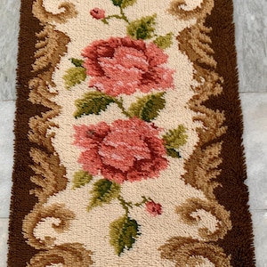 May include: Rectangular rug with a floral design. The rug features two large pink roses with green leaves on a cream-colored background. The border is brown with decorative swirls. The rug is made of a textured material.