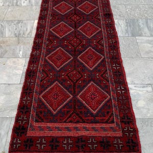 Könnte beinhalten: Ein langer, rechteckiger Läufer mit tiefroter Grundfarbe und einem sich wiederholenden Muster aus dunkelblauen und schwarzen Rauten. Die Bordüre zeigt ein geometrisches Kreuzmotiv und die Enden sind mit einem dunklen Band und Fransen versehen.