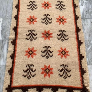 May include: Rectangular beige rug with a repeating pattern of orange and dark brown floral and abstract designs. The rug has a dark brown border with a zig-zag pattern. The rug is made of a textured material.