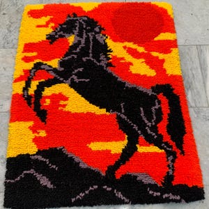 May include: A rectangular rug featuring a black horse rearing against a sunset backdrop. The rug's design incorporates shades of red, yellow, and black, creating a vibrant and dynamic scene. The rug is made of a textured material.