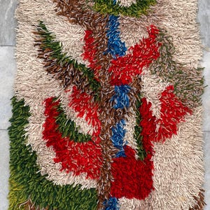 May include: A rectangular, textured rug with a colorful abstract design. The rug features a cream-colored background with a central pattern of red, green, brown, and blue shapes. The rug's design is reminiscent of a stylized floral or landscape motif.