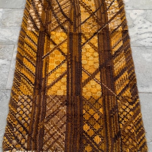 May include: A rectangular area rug featuring a high-pile texture with a geometric pattern in shades of mustard yellow, golden orange, and dark brown. The design includes vertical stripes and diamond motifs, finished with a light beige fringe at the bottom.