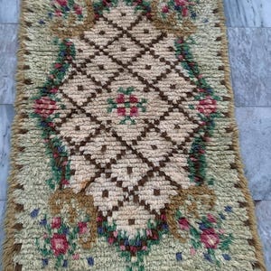 May include: Rectangular rug with a beige diamond pattern center, framed by a border of tan and green. The rug features floral accents in pink, red, and blue. The rug has a shaggy texture and a fringed edge.