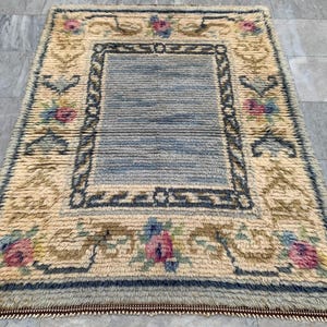 May include: Rectangular rug with a central blue panel and a cream-colored border. The border features floral and scrollwork designs in pink, blue, and brown. The rug has a fringed edge and a textured surface.