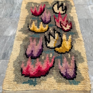 May include: A rectangular rug with a beige border and a gray central field. The rug features a repeating pattern of stylized, flame-like shapes in pink, purple, yellow, and white, outlined in black. The rug has a textured, woven appearance.
