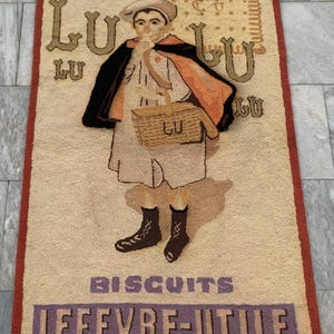 May include: Vintage rug featuring a child with a basket, wearing a cape, and a hat. The rug has the words "LU" and "BISCUITS LEFEVRE-UTILE" in a decorative font. The rug has a cream background with a red border.