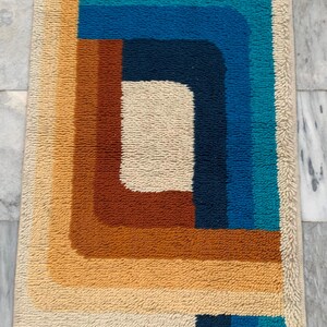 May include: A rectangular, retro-style rug with a geometric design. The rug features a cream-colored border and a central pattern of layered rectangles in shades of brown, orange, blue, and teal. The rug's texture appears soft and plush.