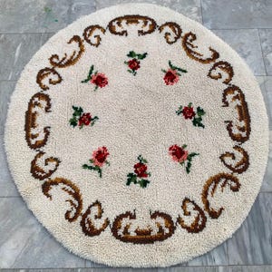 May include: A round, off-white shag rug featuring a circular border of brown scrollwork patterns. The center of the rug is decorated with six small, scattered floral motifs, each consisting of red rose petals and green leaves on a textured surface.