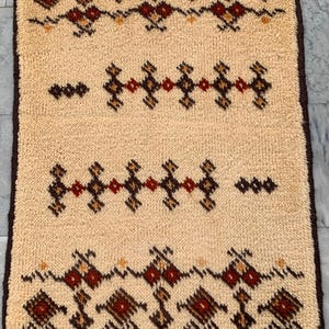 May include: A rectangular rug with a thick, cream-colored pile featuring dark brown, red, and orange geometric diamond patterns arranged in horizontal bands. The rug is finished with a dark brown border along all four edges of the textured fabric.