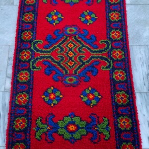 May include: A rectangular rug featuring a vibrant red background with a central blue and green geometric floral pattern. The border consists of a dark blue frame decorated with repeating small red, green, and yellow flower motifs.