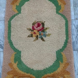 May include: A rectangular tufted rug featuring a beige center with a central floral bouquet of pink, yellow, and blue flowers. The rug is bordered by a green scalloped frame and accented with gold-toned scrollwork on a textured tan background.