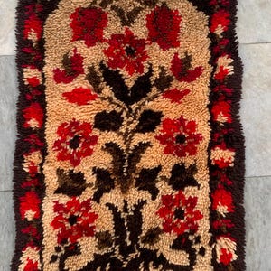 May include: A rectangular shag rug featuring a central beige field with a symmetrical floral pattern in deep red and dark brown. The rug is framed by a dark brown border with red floral accents and finished with dark brown fringe at the bottom.