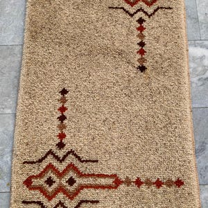 May include: A rectangular beige rug with a thick, textured pile and knotted fringe at both ends. The rug features two symmetrical geometric patterns in shades of dark brown, rust orange, and tan, consisting of jagged lines and diamond shapes.