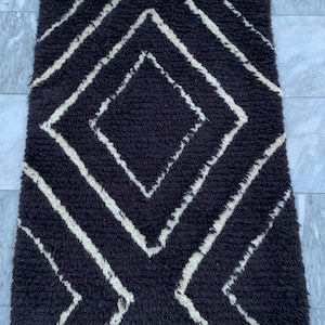May include: A rectangular rug with a thick, dark brown, shaggy pile featuring a contrasting cream-colored geometric pattern. The design consists of a large central diamond shape flanked by smaller angular lines that extend toward the top and bottom edges.