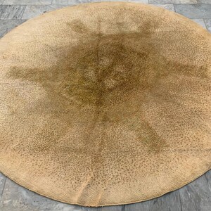 May include: A large, round rug with a light brown, textured surface. The rug has a mottled appearance with darker brown spots and a slightly darker center. The rug is placed on a gray tiled floor.