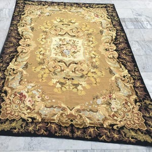May include: A large rectangular rug with an ornate floral design. The rug has a tan centre with a detailed floral pattern in shades of yellow, cream, and red. A dark brown border with leaf motifs frames the design, creating a classic aesthetic.