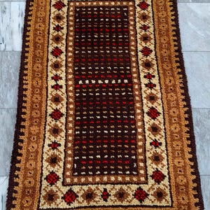 May include: A rectangular rug featuring a deep brown central field with a grid of small red and cream-colored tufted accents. The border consists of a cream-colored band with red and brown geometric motifs, framed by a thick, textured brown edge.
