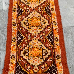 May include: A rectangular, vintage-style rug with a repeating geometric pattern in shades of orange, brown, and cream. The rug has a thick, shaggy texture and a rust-colored border. The design features stylized floral and geometric shapes.