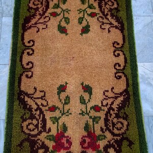 May include: A rectangular rug with a textured pile featuring a tan center panel and a wide olive green border. The design includes dark brown scrollwork accents and symmetrical floral motifs with red roses and green leaves at both ends.
