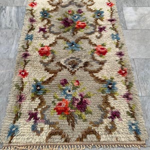 May include: A rectangular, beige rug with a floral pattern. The rug features a central design of flowers in red, blue, and purple, surrounded by a brown border. The rug has a fringed edge and is placed on a gray tiled floor.