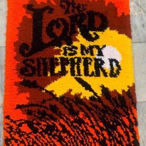 May include: A rectangular, handmade rug with the text "The Lord is my Shepherd" in black, brown, yellow, and white. The rug has an orange border and a textured, tufted appearance. The design features a stylized sun and grassy area.