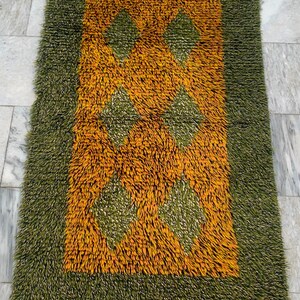 May include: A rectangular rug with a shaggy texture. The rug features a green border and a central design in shades of orange and green. The central design includes diamond shapes. The rug is designed for home decor.