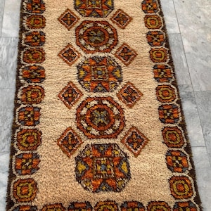 May include: Rectangular rug with a beige central field and a dark brown border. The rug features geometric patterns in shades of orange, yellow, and blue. The design includes repeating circular and diamond shapes.