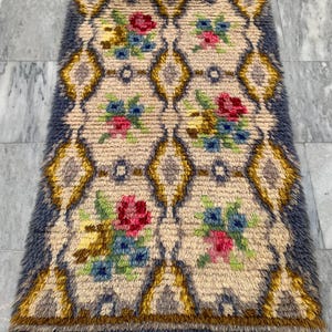 May include: Rectangular rug with a cream-colored background and a repeating diamond pattern. The diamonds are outlined in gold and gray, with floral clusters in red, blue, and yellow. The rug has a blue fringe border.