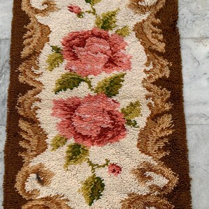 May include: Rectangular rug with a floral design. The rug features two large pink roses with green leaves on a cream background. A brown border with decorative swirls surrounds the floral pattern. The rug is made of a textured material.