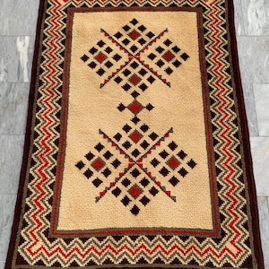 May include: A rectangular rug featuring a beige center with two symmetrical geometric diamond patterns in dark brown and red. The rug is framed by a wide border with a repeating zigzag pattern in dark brown, red, and beige, finished with a solid dark brown edge.