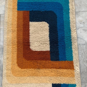May include: A rectangular, retro-style rug with a cream-colored border. The rug features a geometric design with blocks and curved lines in shades of brown, orange, blue, and teal. The rug has a textured, woven appearance.