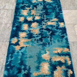 May include: A rectangular rug with a shaggy texture. The rug features a pattern of teal, turquoise, and beige colors. The design has a floral or abstract appearance, with the colors blending together to create a unique visual effect.